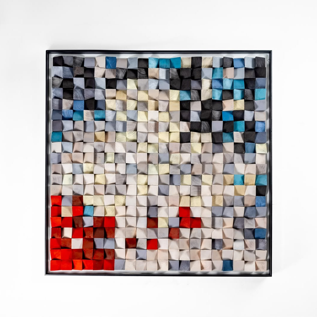 A square framed abstract mosaic artwork made of many small 3D cube-like blocks in black, white, gray, blue, and red, hanging against a plain white wall.